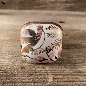 May include: A white ceramic knob with a gold rim and a floral design featuring a bird in pink, gold, and brown.