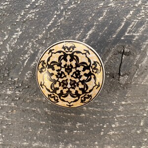 May include: A round, ceramic knob with a black and white floral design. The knob has a cream background and black floral accents.