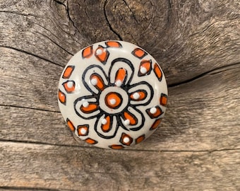 May include: A round ceramic knob with a white background and a black and orange floral design. The knob has a raised, circular center with a white dot in the middle.