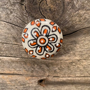 May include: A round ceramic knob with a white background and a black and orange floral design. The knob has a raised, circular center with a white dot in the middle.