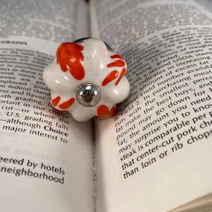 Pode incluir: Um botão de cerâmica branca com um acento prateado e um padrão floral laranja. O botão é um elemento decorativo para móveis ou armários.