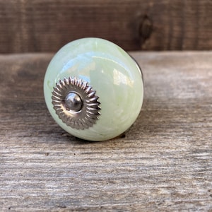 Może przedstawiać: Zielony ceramiczny gałka z srebrnym metalowym akcentem. Gałka ma wzór wiru i idealnie nadaje się do dodania odrobiny elegancji do każdego pomieszczenia.