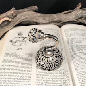Pode incluir: Um gancho de cerâmica branca com um design floral preto e branco. O gancho está preso a uma base de metal prateado. O gancho está em cima de um livro sobre carne.