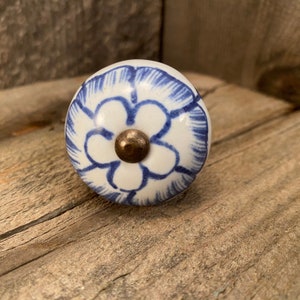 May include: A white ceramic knob with a blue floral design. The knob has a gold metal center.