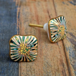 May include: Two square ceramic cabinet knobs with a floral design. The knobs are white with green and gold accents. The floral design features a yellow center with green petals and gold accents.