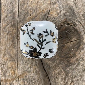 Op de afbeelding: Witte keramische knop met een zwart en gouden bloemenontwerp.