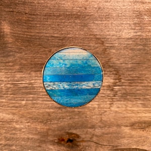 Puede incluir: Anillo de forma ovalada con una incrustación rayada azul y blanca. El anillo es de metal y tiene un acabado pulido.