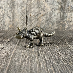 May include: A small, metal, bronze-coloured triceratops figurine. The figurine is detailed with textured scales and has a long tail.