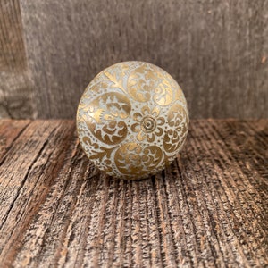 May include: A spherical cabinet knob with a distressed white base and gold floral patterns. The metal knob is decorated with intricate designs and sits on a weathered wooden surface.