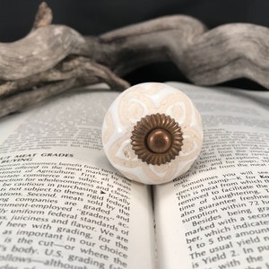 May include: A white and beige ceramic cabinet knob with a floral design and a bronze-colored center. The knob is round and sits on an open book. The knob is a decorative element for furniture and home decor.