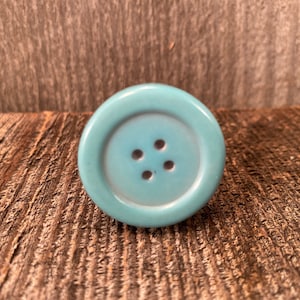 以下が含まれることがあります： 4つの穴のある薄い青色のセラミックボタン。ボタンは丸く、縁が盛り上がっています。