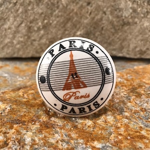 May include: White ceramic knob with a black and brown Eiffel Tower design. The knob has the word "Paris" written around the edge in black.