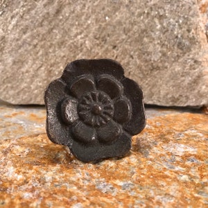 Könnte beinhalten: Ein dunkler Metall-Blumenknauf im antiken Stil. Der Knauf zeigt ein detailliertes, dreidimensionales Rosendesign mit mehreren Blütenblättern und einem zentralen Blumenmuster. Der Knauf ist vor einem natürlichen Steinhintergrund platziert.