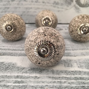Op de afbeelding: Vier witte keramische kastknoppen met een gekrakelde afwerking en zilveren metalen accenten. De knoppen zijn rond en hebben een reliëf in het midden.