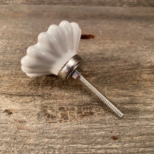 Pu&ograve; includere: Pomello per armadio in ceramica bianca con design scanalato. Il pomello ha una base in metallo argentato e una vite filettata per l'installazione. L'hardware &egrave; posto su una superficie di legno stagionato.