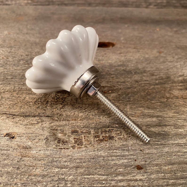 Pu&ograve; includere: Pomello per armadio in ceramica bianca con design scanalato. Il pomello ha una base in metallo argentato e una vite filettata per l'installazione. L'hardware &egrave; posto su una superficie di legno stagionato.