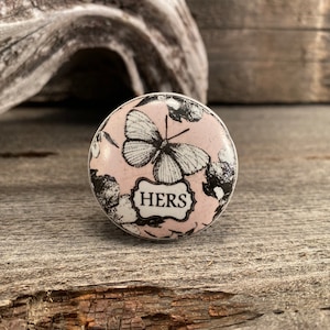 May include: A round, white ceramic knob with a pink background and a black and white butterfly design. The knob has the word "HERS" written in black on a white banner.
