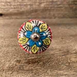 Peut inclure: Un bouton en céramique avec un motif floral bleu, jaune et blanc. Le bouton a une bordure rouge et un centre en métal argenté.