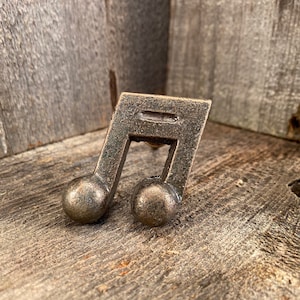 May include: A bronze-colored musical eighth note cabinet knob. The knob has a rectangular top with a horizontal line and two round spheres at the bottom. The knob is set against a weathered wood background.