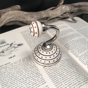 May include: A white ceramic hook with brown polka dots and a brown and white striped knob. The hook is mounted on a silver base and is ideal for hanging items in a bathroom or kitchen.