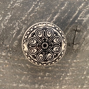 May include: A white ceramic knob with a black and white geometric design. The design features a central circle with a repeating pattern of teardrop shapes and stars.