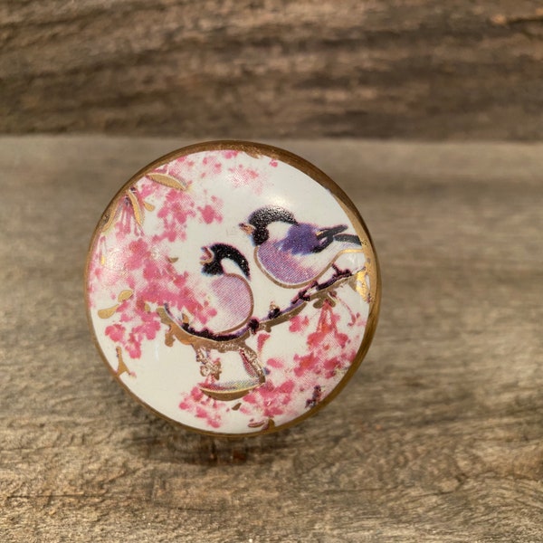 Könnte beinhalten: Ein weißer Keramik-Knopf mit goldfarbenem Rand und einem floralen Design mit zwei rosa Vögeln auf einem Zweig mit rosa Blüten.