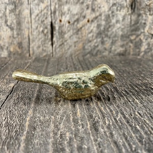 May include: A gold-coloured metal bird figurine with a textured surface. The bird is facing to the right and has a long, thin beak.
