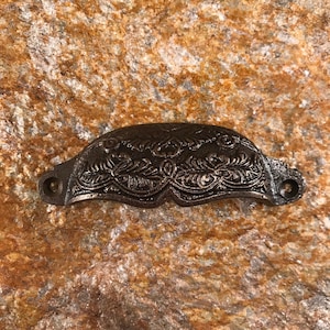 Puede incluir: Un tirador de cajón de metal oscuro y ornamentado con un diseño floral detallado. El tirador es curvo con dos agujeros para el montaje. El acabado tiene un aspecto rústico y envejecido, lo que sugiere un estilo vintage o antiguo.