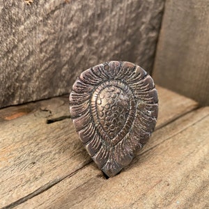 May include: A detailed, antique-style metal ring with a teardrop shape. The ring features an intricate, embossed design with a central textured oval and surrounding floral-like patterns. The ring has a tarnished, aged appearance.
