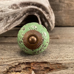 May include: A green ceramic knob with a distressed finish and a brass-colored center. The knob has a raised, circular design around the center.