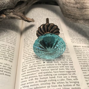 Pode incluir: Um puxador decorativo para móveis com um topo de vidro facetado azul-claro translúcido com um padrão radial. A base do puxador é feita de metal escuro com tom de bronze antigo e um design estriado e festonado ligado a um pino central.