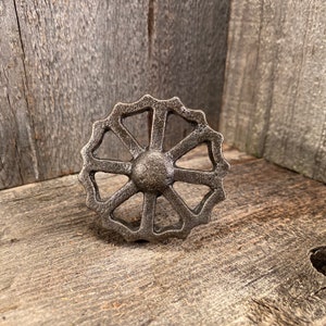 Könnte beinhalten: Ein gusseiserner Knopf in Radform mit einem runden Zentrum und acht Speichen. Der Knopf ist dunkelgrau und hat eine rustikale Oberfläche.