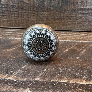 May include: A decorative cabinet knob with a gold-colored base and a round, patterned top. The top features a black and white floral design with a gold center. The knob is set against a rustic, wooden background.