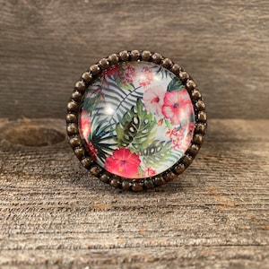 May include: A round, floral patterned cabinet knob with a bronze metal frame. The knob features a white background with pink and green tropical flowers and leaves.