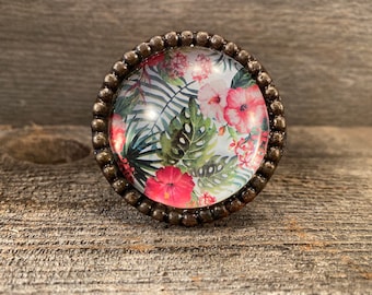 Pode incluir: Um botão de armário redondo com padrão floral e moldura de metal bronze. O botão apresenta um fundo branco com flores e folhas tropicais rosa e verdes.