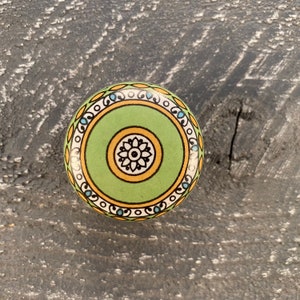 May include: A round, decorative cabinet knob with a green center and a floral design. The knob features a white, yellow, and blue patterned border. The knob is set against a dark, textured wooden surface.