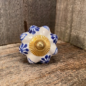 Op de afbeelding: Witte keramische knop met een blauw bloemenpatroon en een gouden midden. De knop heeft een gegolfde rand.