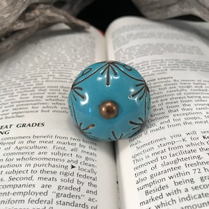 Peut inclure: Un bouton en céramique bleu turquoise avec un motif géométrique noir et blanc. Le bouton a un centre doré.