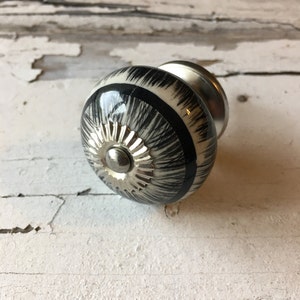 以下が含まれることがあります： 銀色の金属製のベースに、黒と白のセラミック製のノブ。ノブはストライプ模様で、装飾的な銀色のセンターがあります。