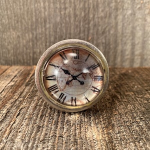 May include: A silver-toned knob with a round, glass face featuring a vintage clock design with Roman numerals. The clock face has a distressed, aged look with a brown and white background.