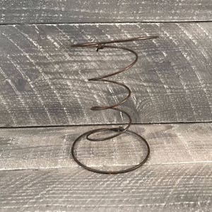 May include: A rusty metal spring coil, commonly used as a base for a bed, is shown against a gray wooden background. The spring is coiled in a spiral shape and has a circular base.