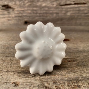 May include: A white ceramic daisy-shaped cabinet knob. The knob has a glossy finish and a detailed, textured center. The daisy petals are evenly spaced around the center. The knob is designed for furniture and home decor.