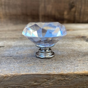 May include: A clear, faceted crystal cabinet knob with a silver-colored base. The knob is diamond-shaped and sits on a tiered, circular base. The background is a weathered, gray wooden surface.