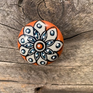 May include: A round ceramic knob with a white and blue floral design on an orange background. The knob has a white center with a blue ring around it.