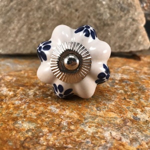 Könnte beinhalten: Ein weißer Keramik-Schrankknopf mit einem blauen Blumenmuster. Der Knopf hat einen gewellten Rand und eine silberne Metallmitte. Der Knopf hat einen Durchmesser von etwa 3,8 cm.