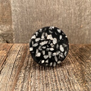 May include: A circular bottle stopper in black, featuring a speckled pattern of white and grey fragments. The stopper is made from a solid material with a smooth, polished finish. Designed for sealing bottles.