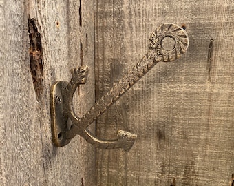 Può includere: Un gancio a forma di ancora in metallo dorato montato su una superficie in legno. L'ancora ha un design decorativo a corda e un anello nella parte superiore.