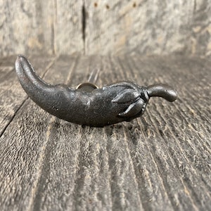 Puede incluir: Un tirador de armario de hierro fundido con forma de chile. El chile es de color gris oscuro con una superficie texturizada y un tallo y hojas detallados. El herraje está montado sobre una superficie de madera envejecida.