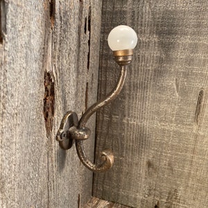Puede incluir: Un gancho de pared con un acabado metálico oscuro, texturizado y de estilo antiguo con un diseño curvo. El extremo superior del gancho está rematado con un pomo de vidrio blanco opaco, liso y redondo, asegurado por un pequeño collar de metal.