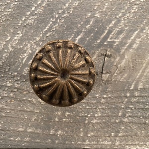 Puede incluir: Primer plano de un pomo de metal dorado con un diseño decorativo en forma de sol. El pomo está colocado sobre un fondo gris claro texturizado.